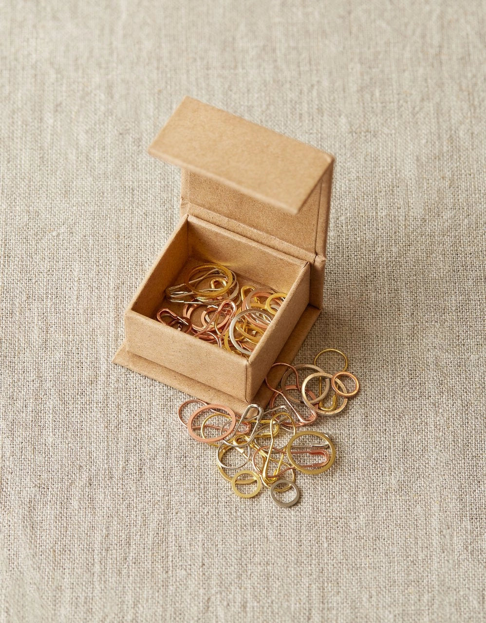 Small open cardboard box with gold and silver stitch markers on a beige fabric background