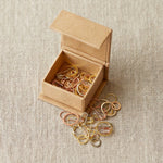 Small open cardboard box with gold and silver stitch markers on a beige fabric background