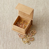 Small open cardboard box with gold and silver stitch markers on a beige fabric background