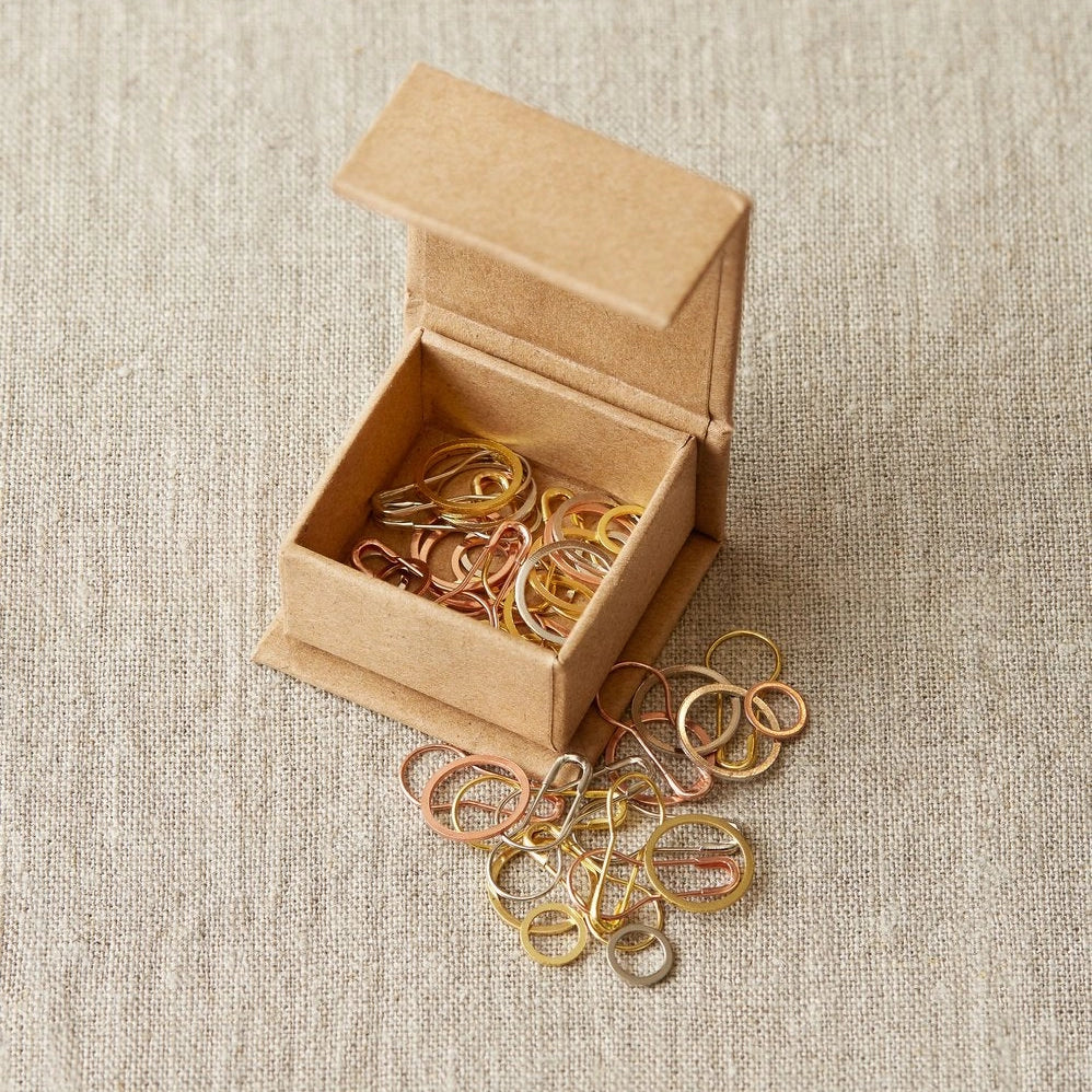 Small open cardboard box with gold and silver stitch markers on a beige fabric background