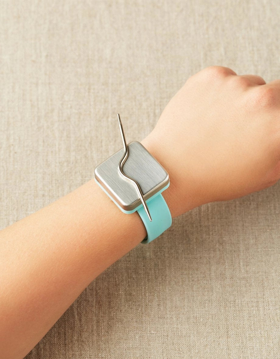Light blue notions holder bracelet with a magnet featured on a person's wrist, with a cable needle attatched to the magnet.