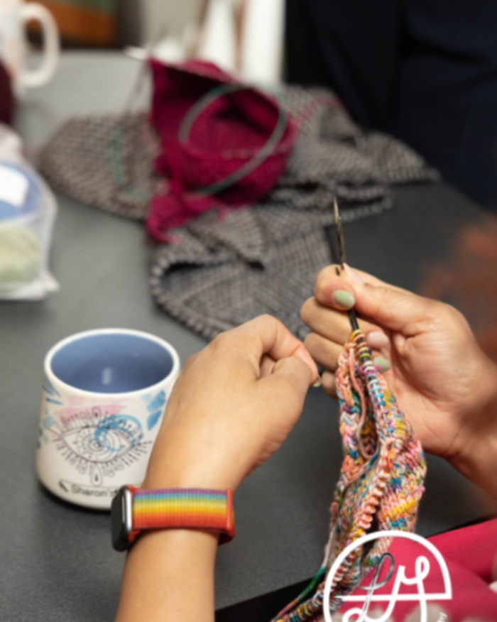 Person knitting with colorful yarn on a table with a mug and yarn ball in the background.