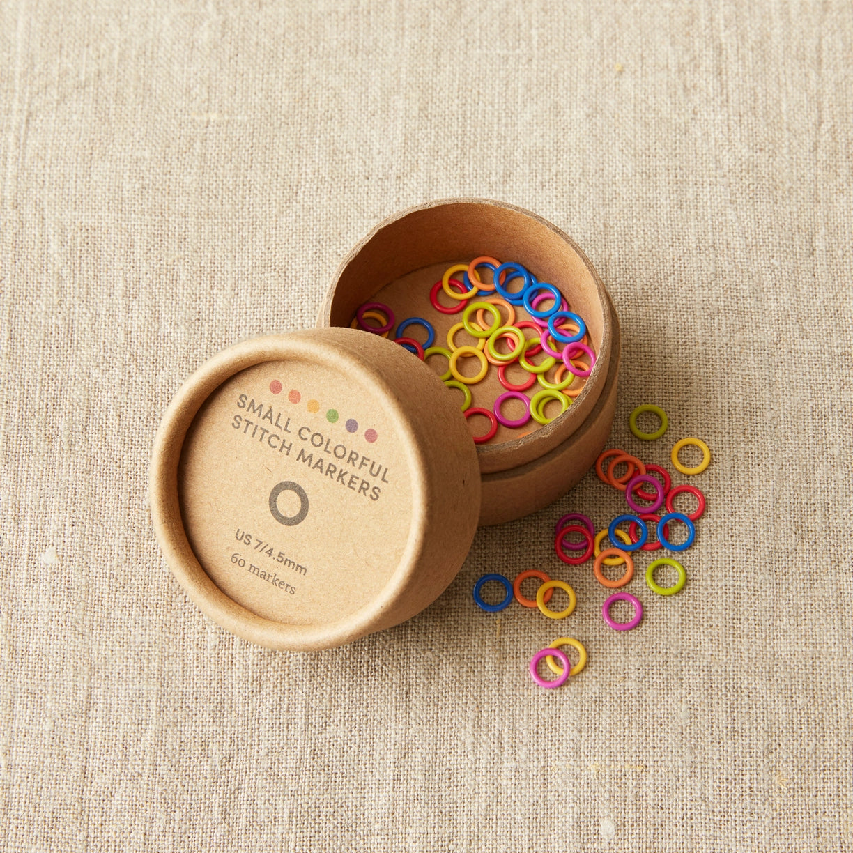 Colorful stitch markers in a wooden container on a beige fabric background