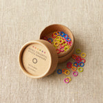 Colorful stitch markers in a wooden container on a beige fabric background
