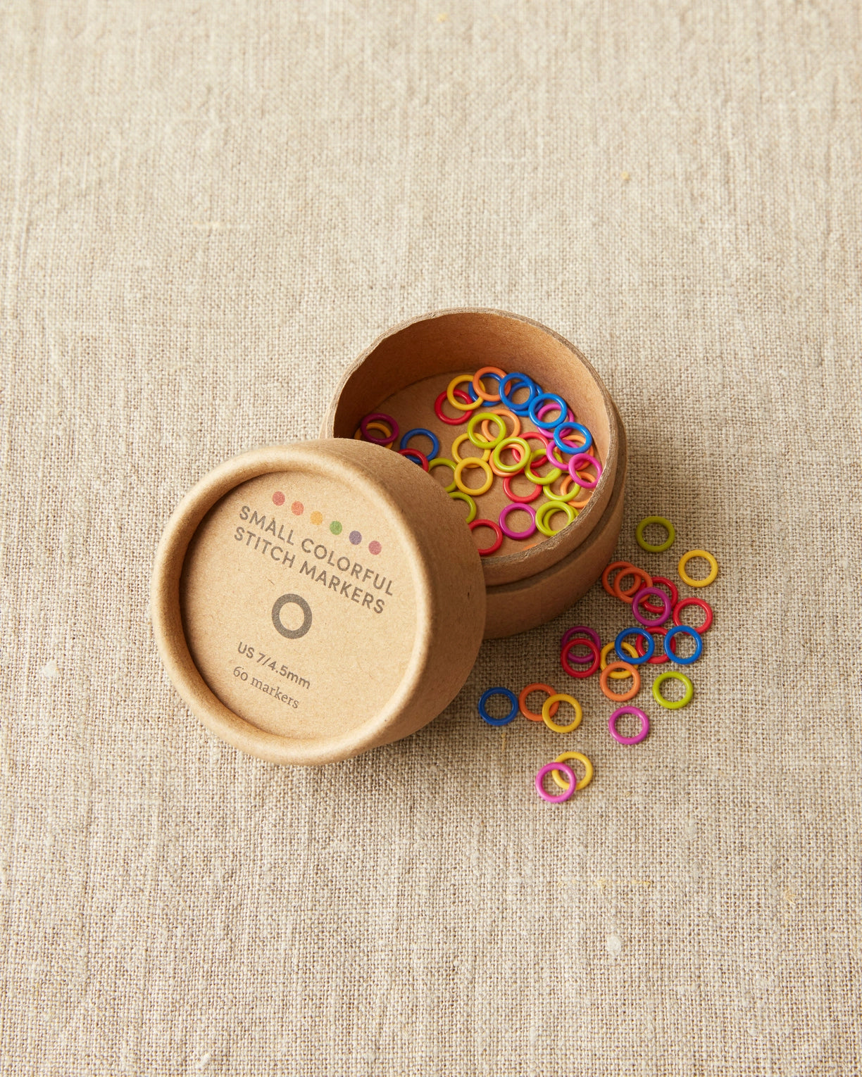 Colorful stitch markers in a wooden container on a beige fabric background