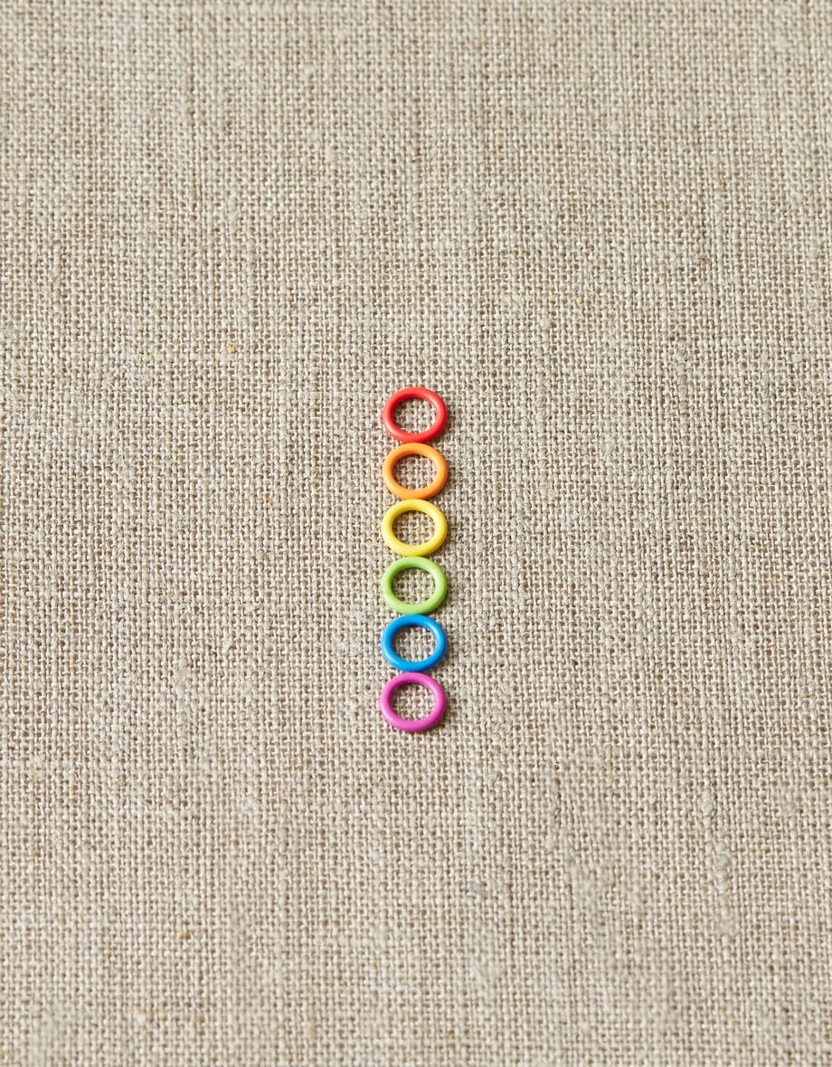 Colorful small ring stitch markers arranged in a vertical line on a beige fabric background