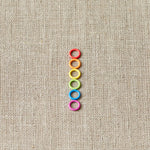 Colorful small ring stitch markers arranged in a vertical line on a beige fabric background