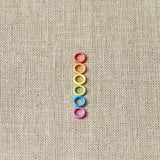Colorful small ring stitch markers arranged in a vertical line on a beige fabric background