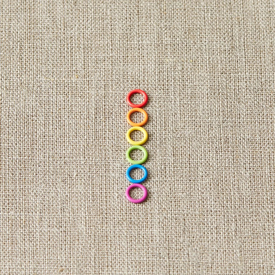 Colorful small ring stitch markers arranged in a vertical line on a beige fabric background