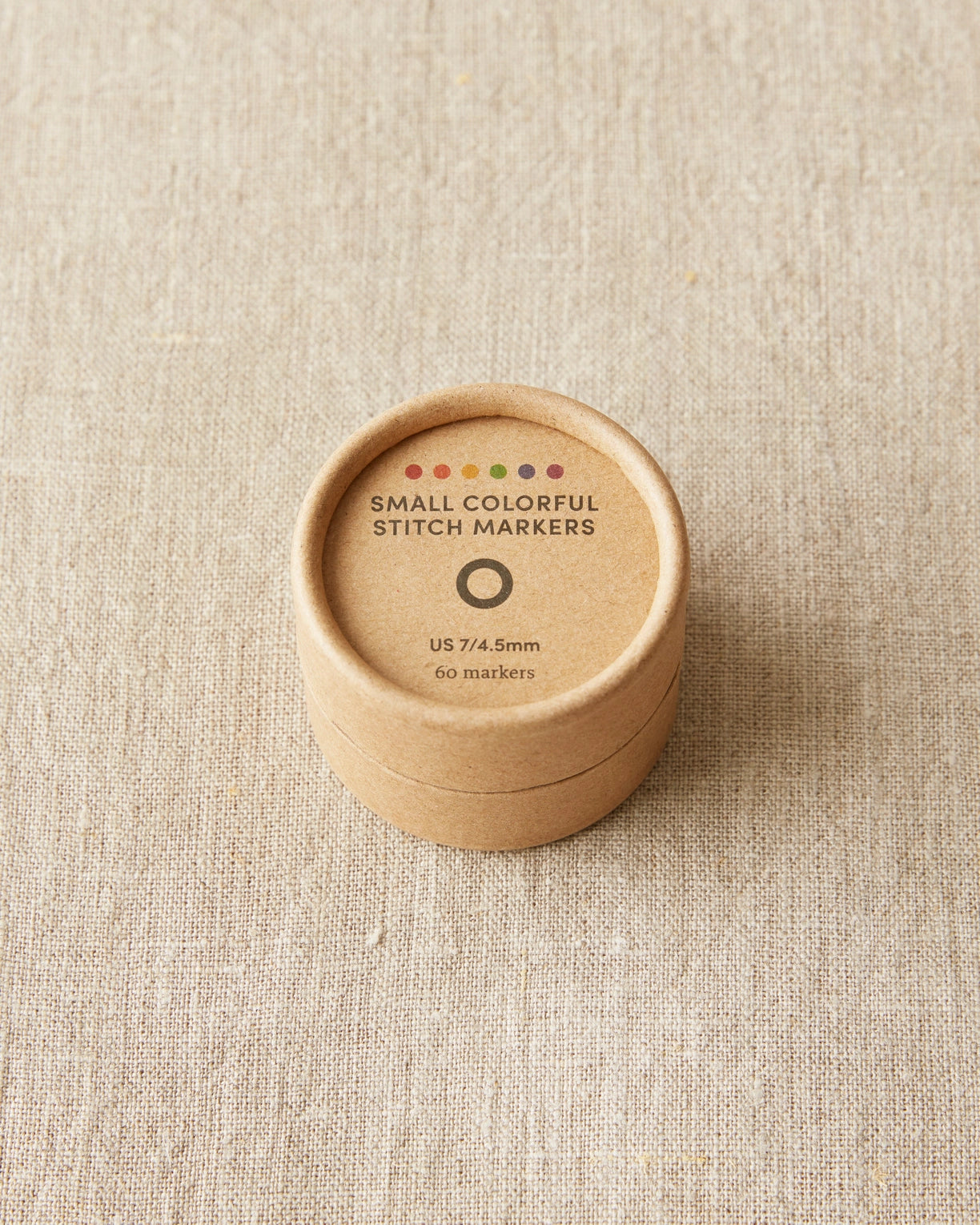 Round brown container with 'Small Colorful Stitch Markers' text on a beige fabric background