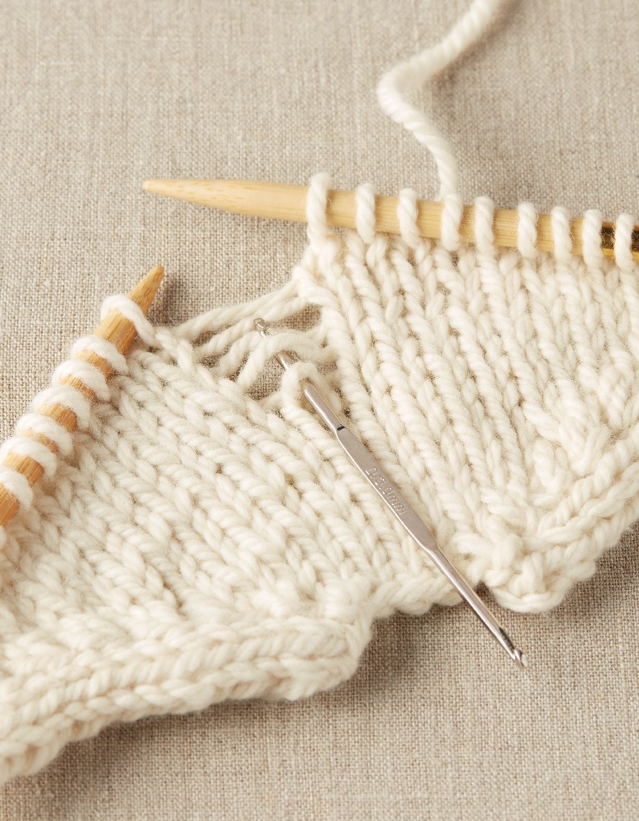 Knitted fabric with wooden knitting needles and a metal crochet hook fixing dropped stitches on a beige background