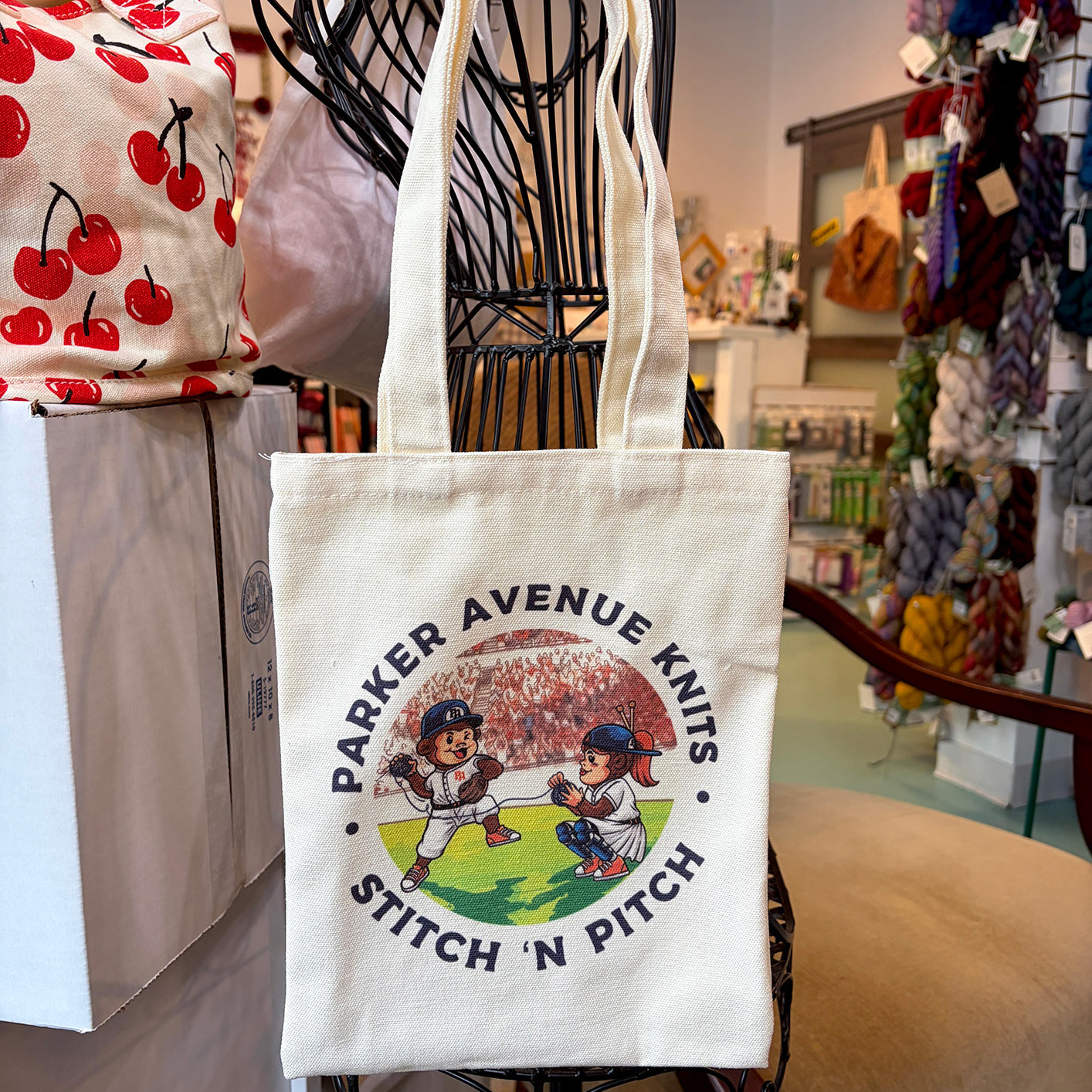 A cream canvas tote bag with a colorful graphic of two baseball players and the text 'PARKER AVENUE KNITS STITCH 'N PITCH' hanging on a black wire display stand inside a yarn shop.