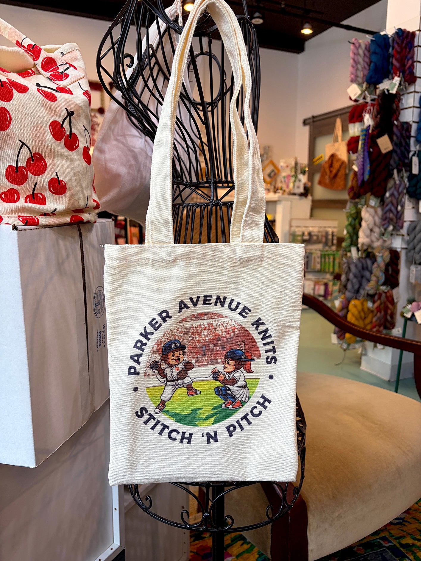 A cream canvas tote bag with a colorful graphic of two baseball players and the text 'PARKER AVENUE KNITS STITCH 'N PITCH' hanging on a black wire display stand inside a yarn shop.