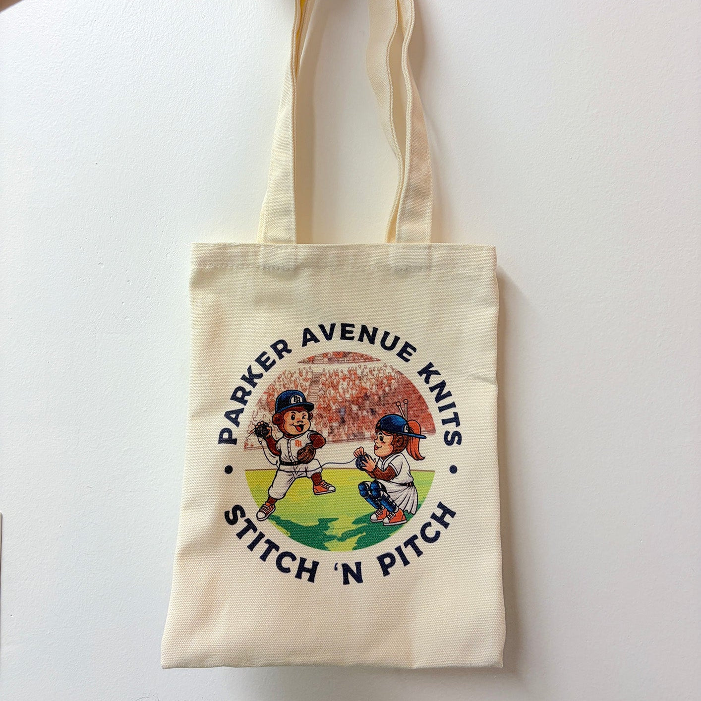 An off-white canvas tote bag with two shoulder straps, featuring a circular graphic print of two cartoon baseball players and the text 'PARKER AVENUE KNITS STITCH 'N PITCH'.