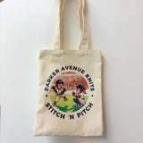 An off-white canvas tote bag with two shoulder straps, featuring a circular graphic print of two cartoon baseball players and the text 'PARKER AVENUE KNITS STITCH 'N PITCH'.