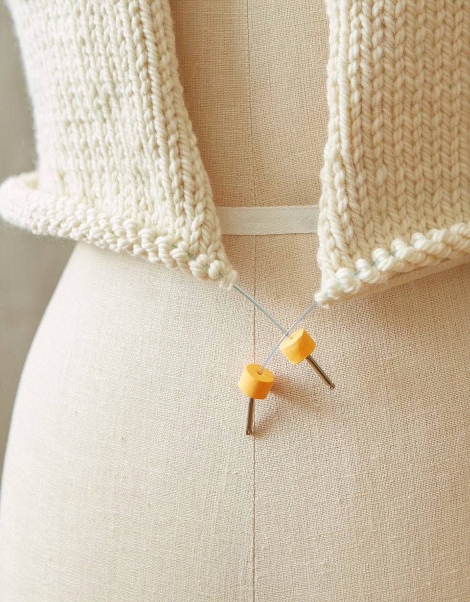 Knitted garment on a mannequin with orange stitch stoppers on circular needles.