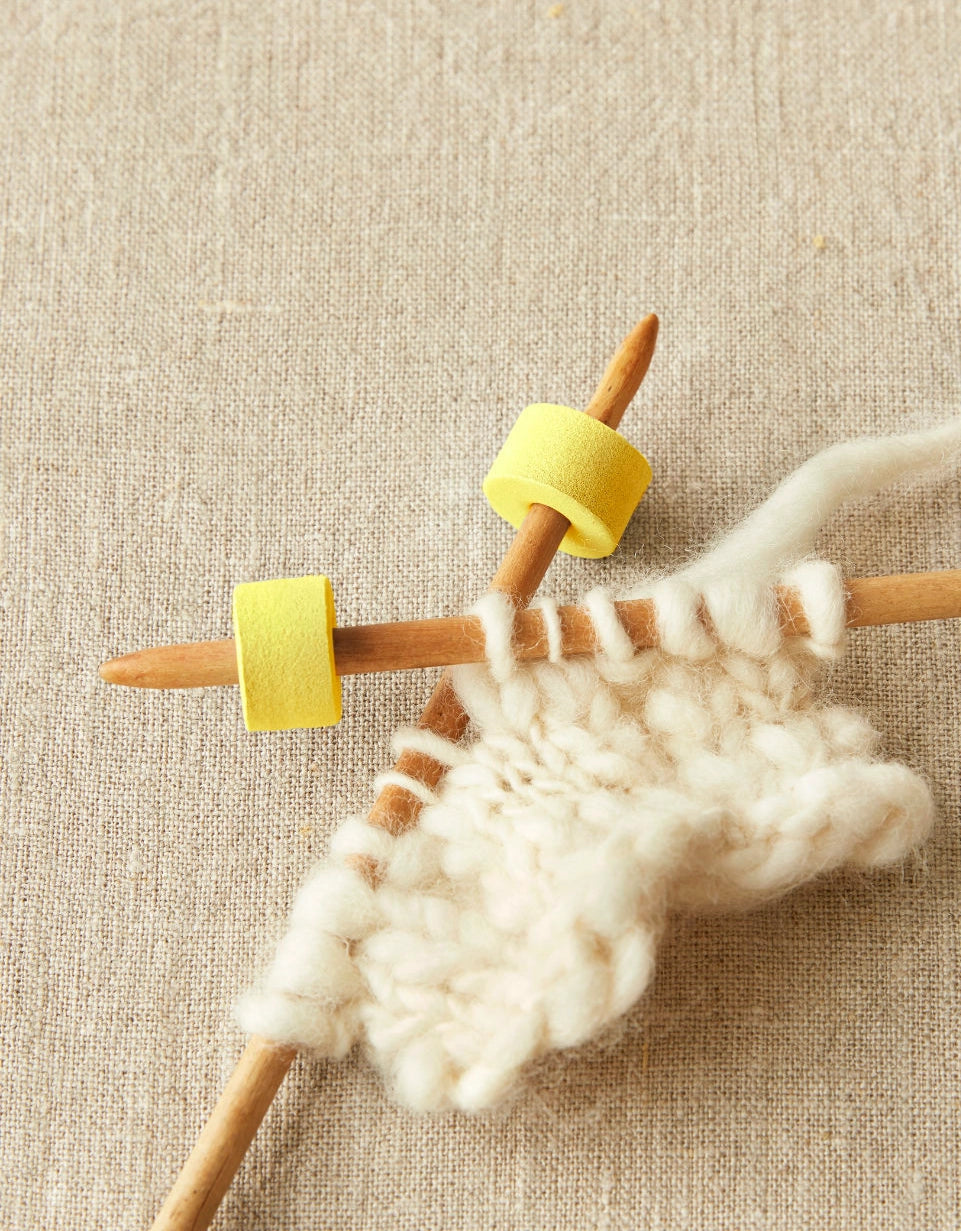 Knitting needles with yellow foam stitch stoppers on a beige fabric background