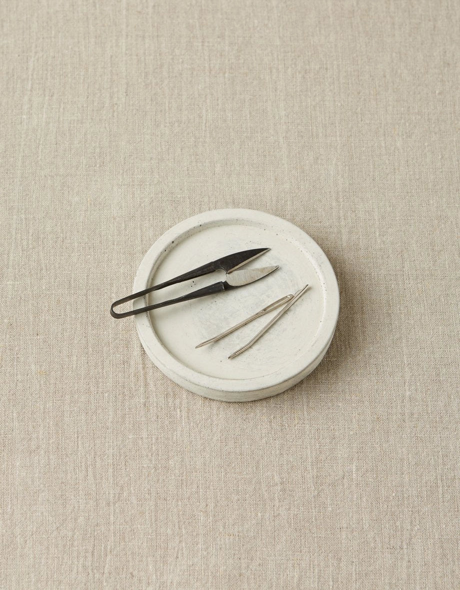 Notions dish with yarn snips and tapestry needles on a beige textured surface