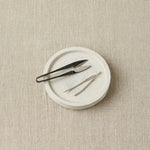 Notions dish with yarn snips and tapestry needles on a beige textured surface