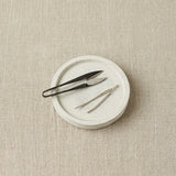 Notions dish with yarn snips and tapestry needles on a beige textured surface