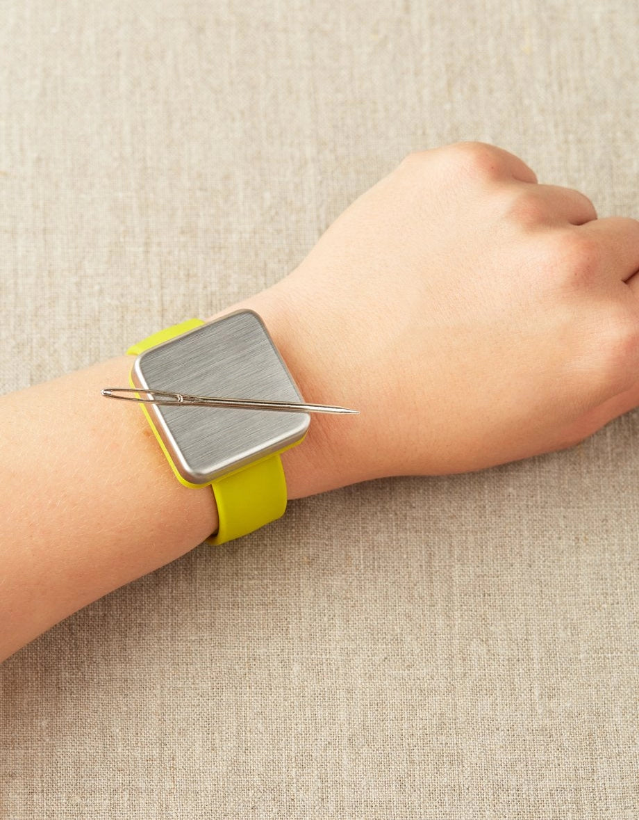 Magnetic slap bracelet with a yellow strap on a wrist with a tapestry needle attatched to the magnet against a beige background