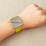 Magnetic slap bracelet with a yellow strap on a wrist with a tapestry needle attatched to the magnet against a beige background