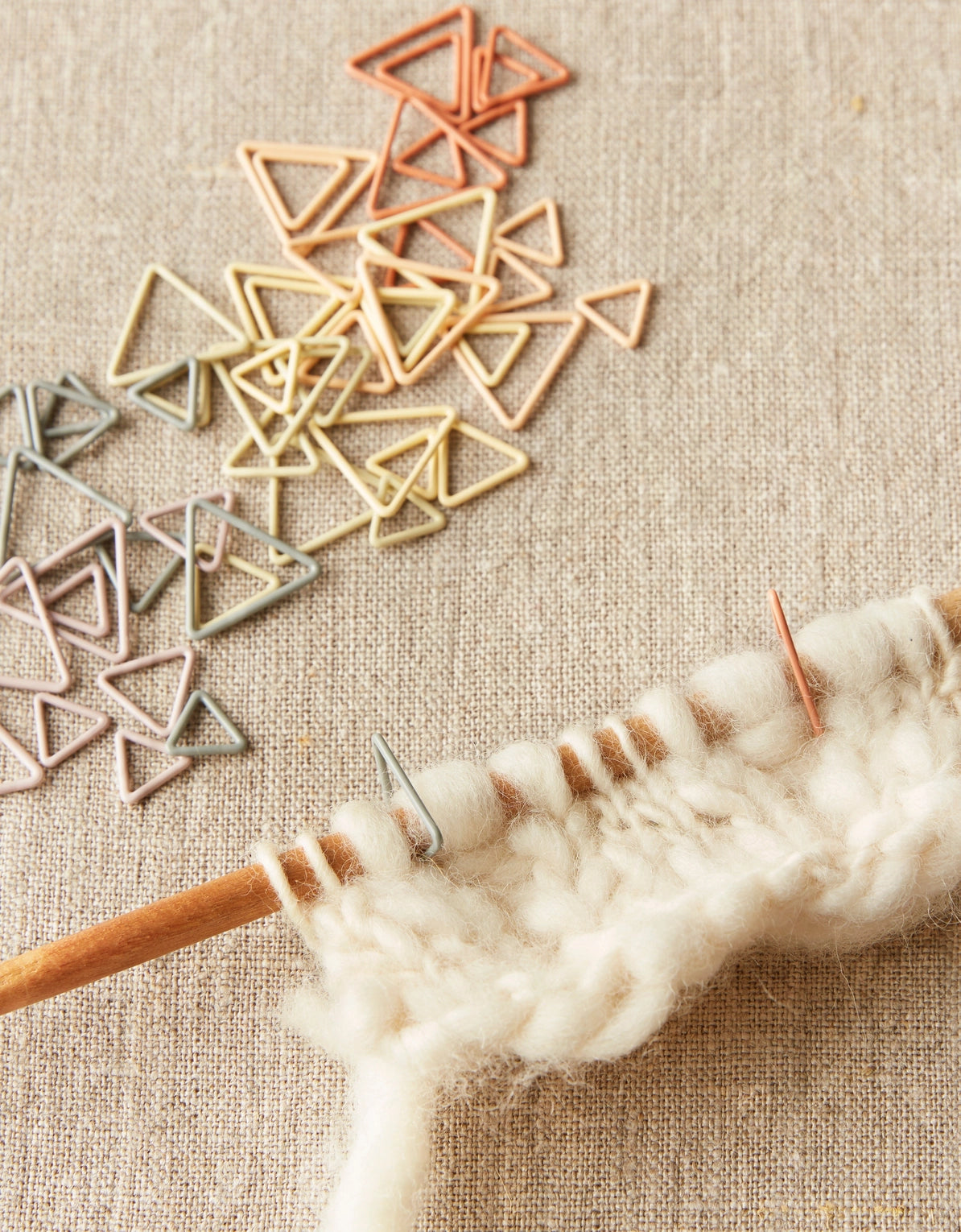 Wooden knitting needle with yarn and triangle shaped stitch markers on the needle on a textured fabric background