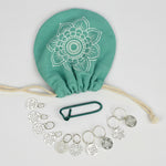 A set of stainless steel knitting needles, a knitting bag with a floral pattern, and a needle threader, all arranged on a white surface.