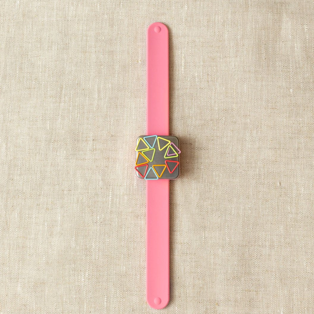 A pink silicone slap bracelet with a brushed steel-plated magnet in the center, holding geometric stitch markers.