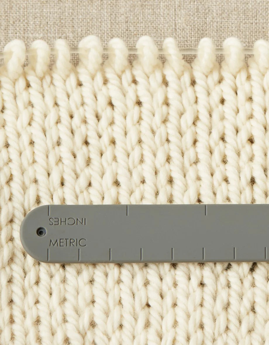 The back of a notions holder bracelet with imperial and metric system measurements, measuring a knitted swatch.